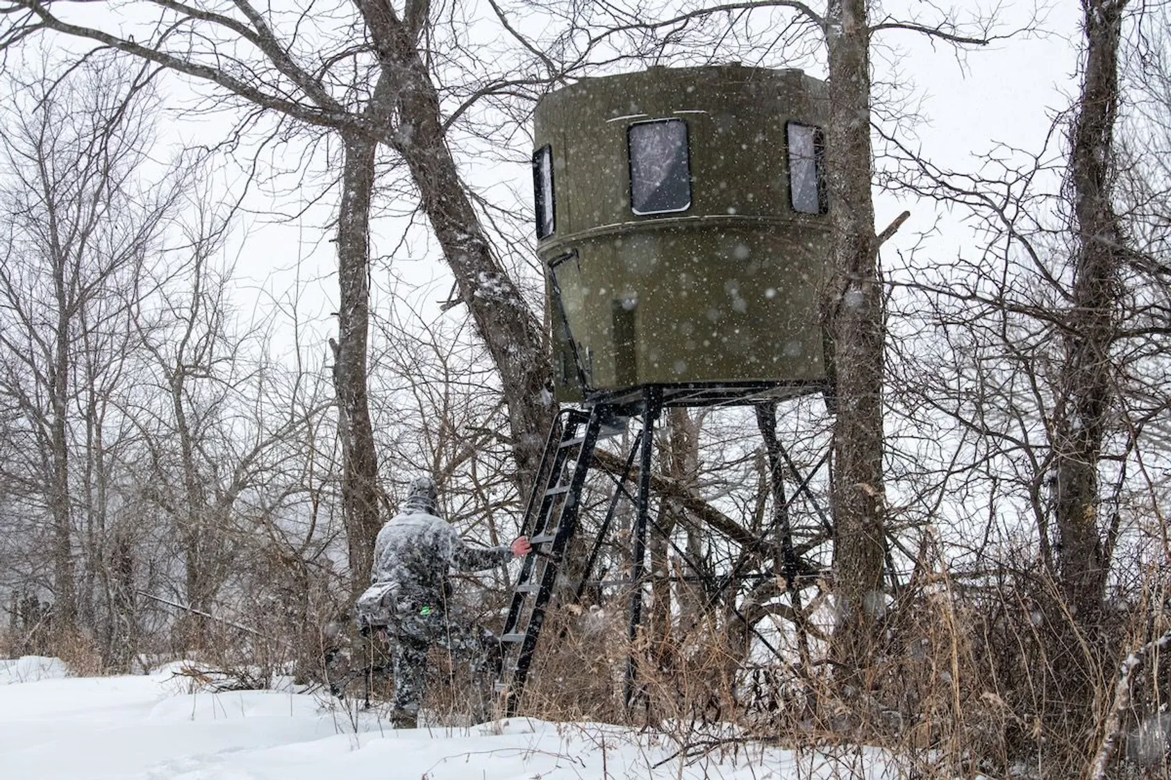 Elevated hunting blind with windows on metal tower stand among bare snow-dusted trees during winter snowfall