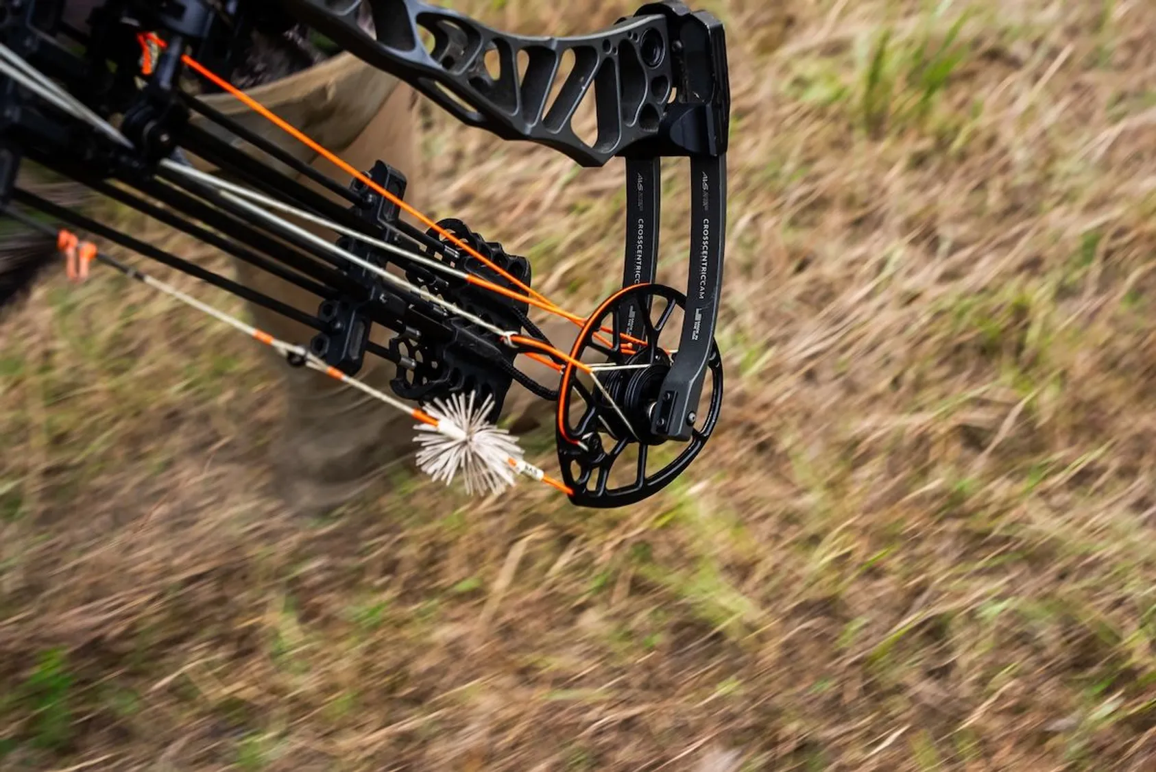 Close-up of black compound bow cam and orange bowstring with white arrow fletching against blurred natural background