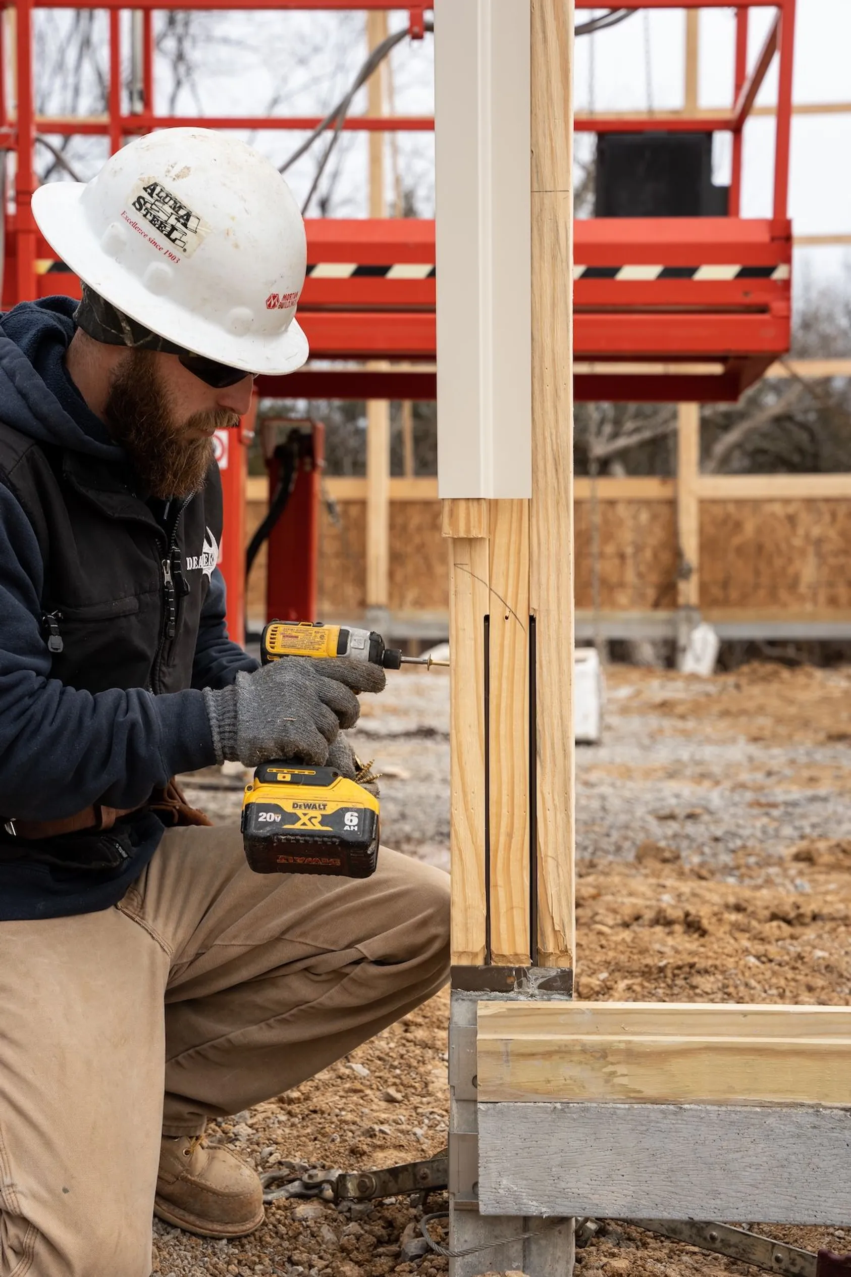 Construction worker in white hard hat and navy jacket using yellow power drill on wooden framing at building site