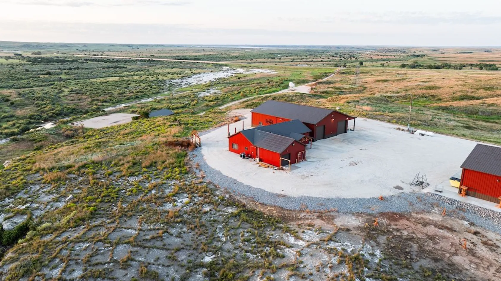Aerial view of red metal buildings on gravel pad with winding stream and mixed grassland near distant horizon