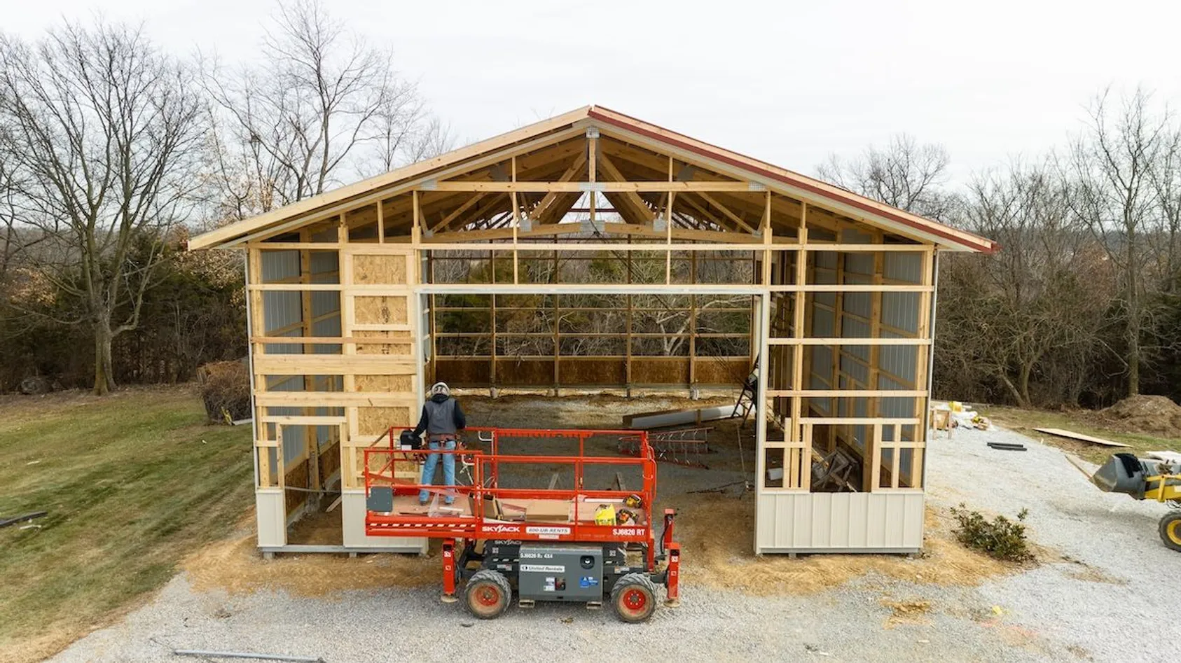 Open-frame pole barn under construction with exposed wood trusses, orange scissor lift, and bare trees in background