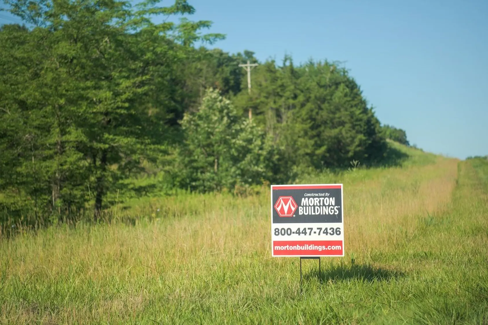Morton Buildings construction sign displaying company logo and phone number in grassy field with dense tree line background