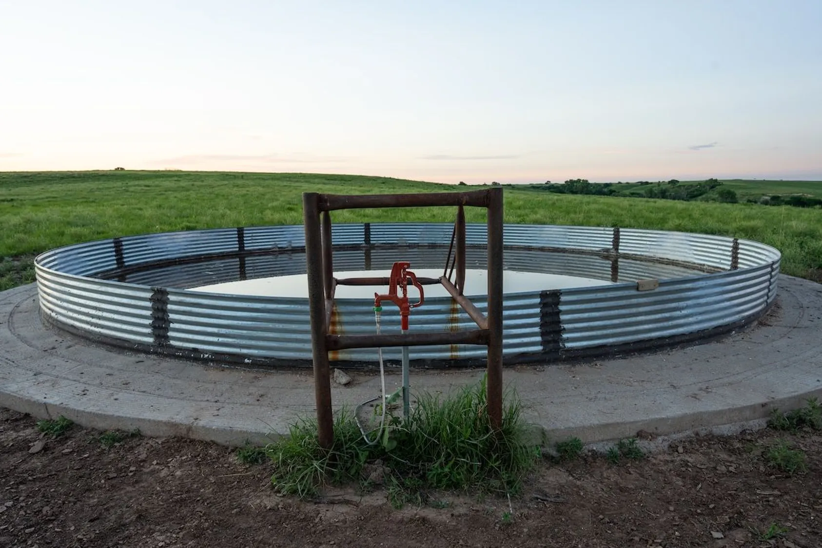 Large circular corrugated metal water storage tank on concrete pad with red pump stand in green prairie landscape.