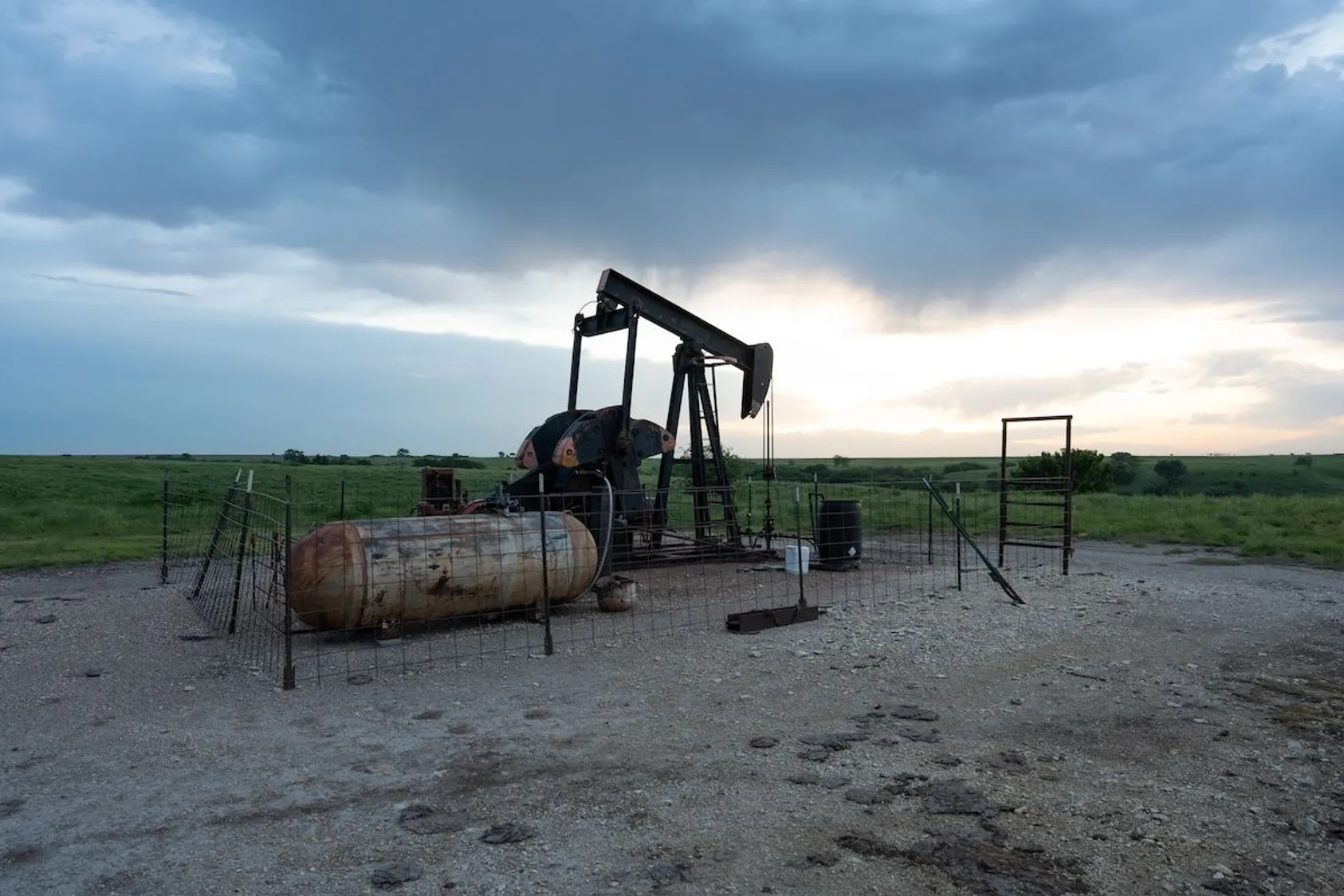 Oil pump jack with storage tanks behind wire fence on gravel pad at dusk with green fields beyond.