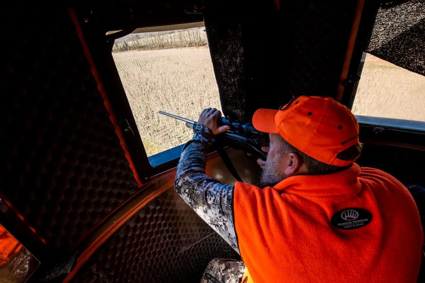 Hunter in orange cap and camouflage aiming scoped rifle out window of elevated hunting blind.