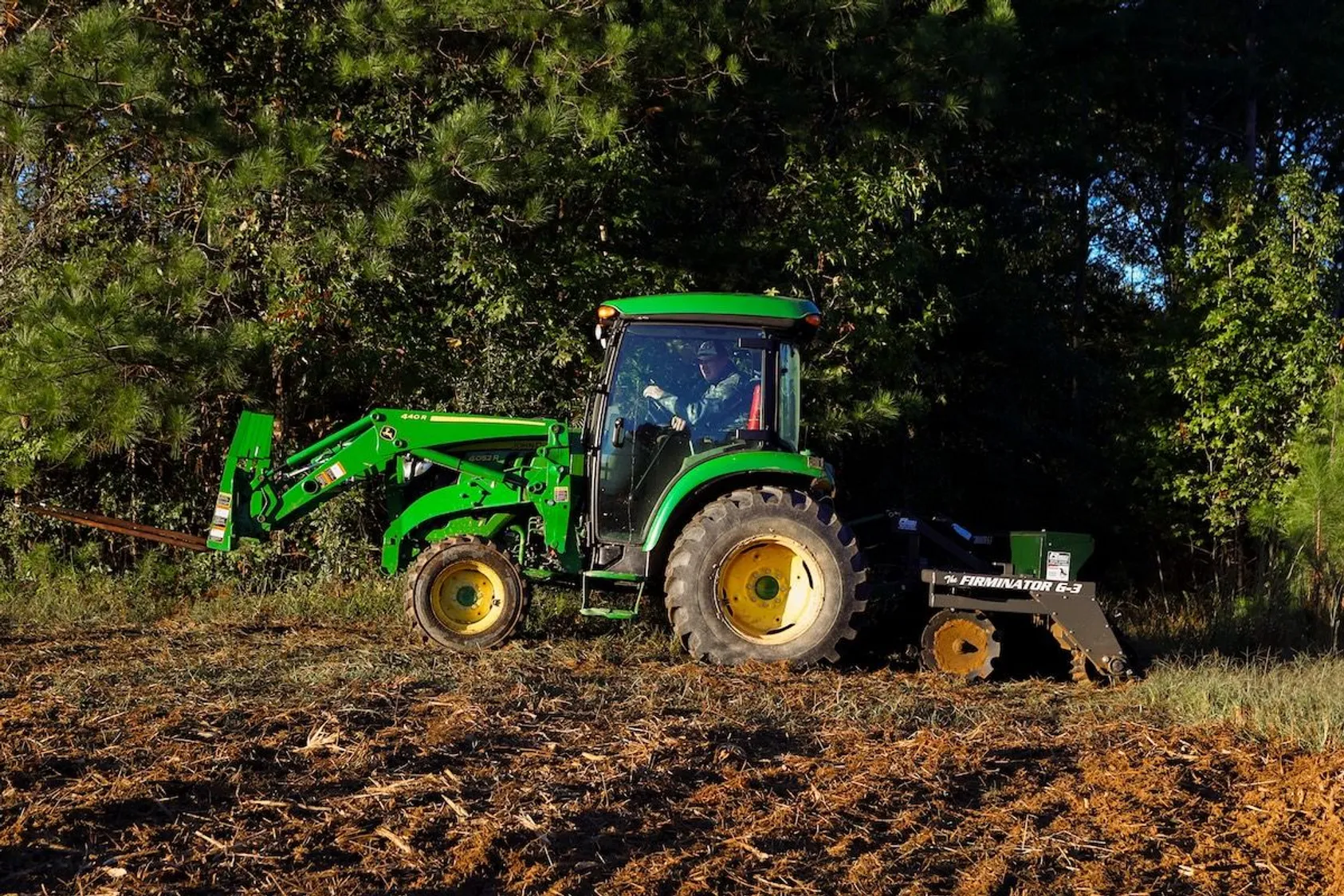 Green tractor with front loader attachment working on cleared brown soil near a wooded area