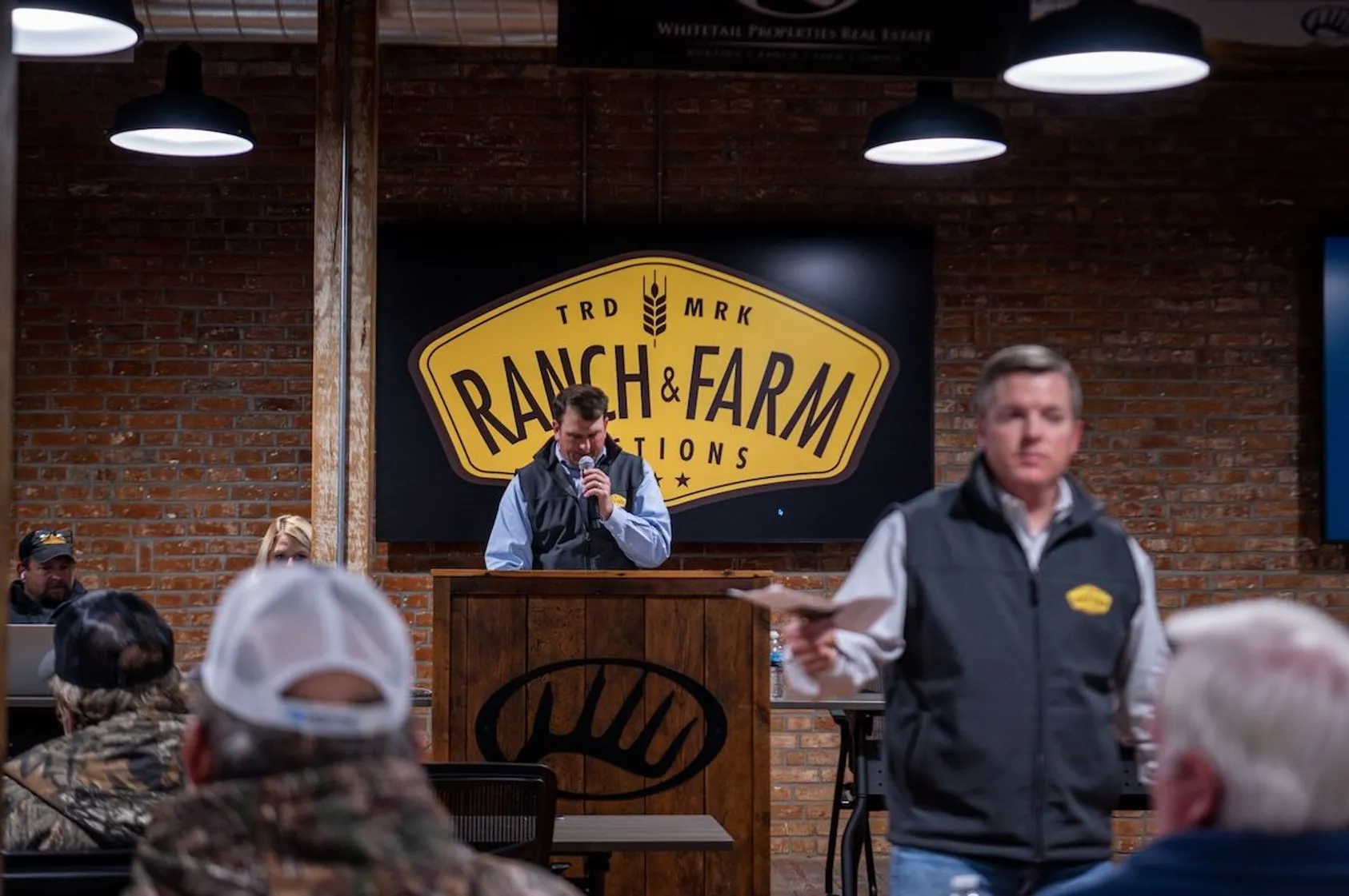 Auctioneer speaking into microphone at podium beneath Ranch & Farm Auctions sign in brick-walled venue with attendees seated