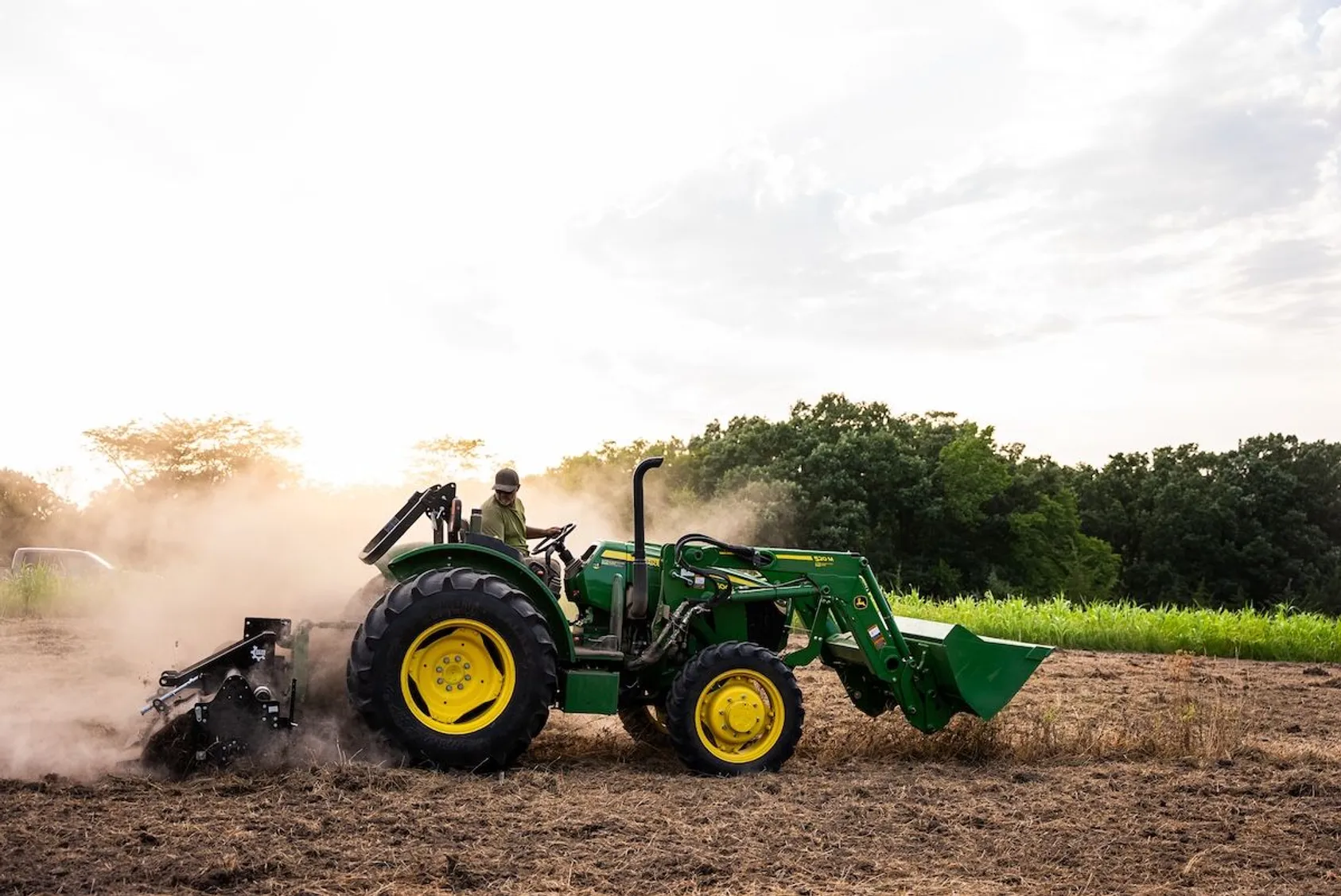 Green John Deere tractor with front loader kicking up dust while tilling soil at sunset near trees