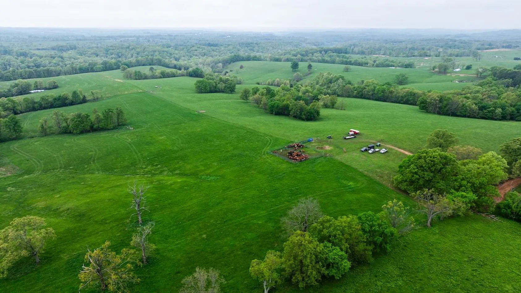 Aerial view of agricultural land with green fields, farm equipment and vehicles gathered in one section, surrounded by wooded areas.