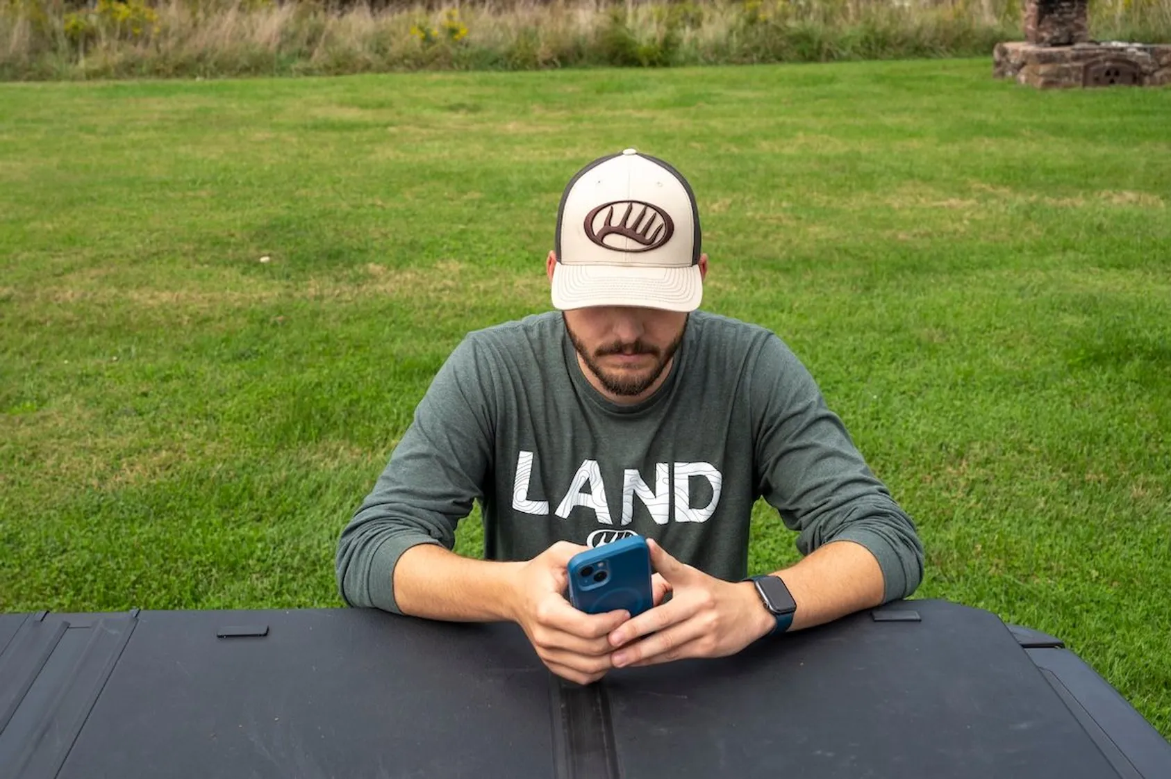 Person wearing a LAND shirt and baseball cap looking down at their phone while sitting at an outdoor table on a lawn.