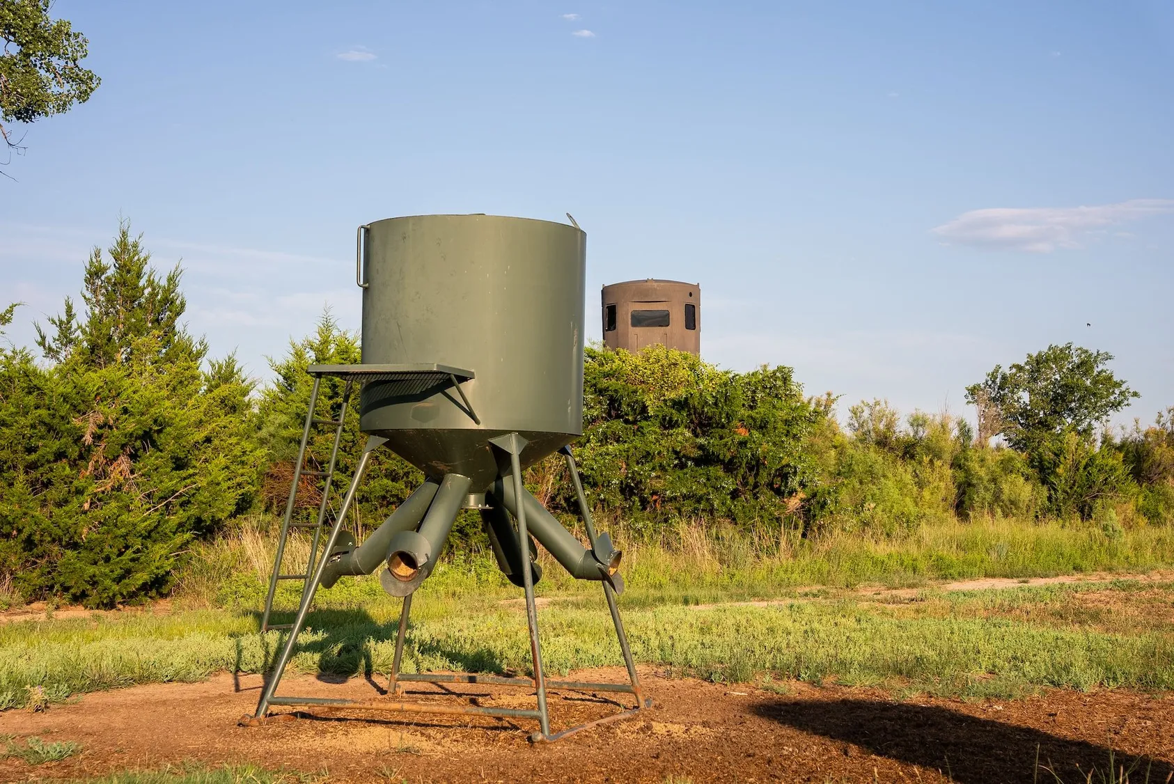 Green cylindrical gravity deer feeder on elevated stand with brown hunting blind in background near trees and green grass