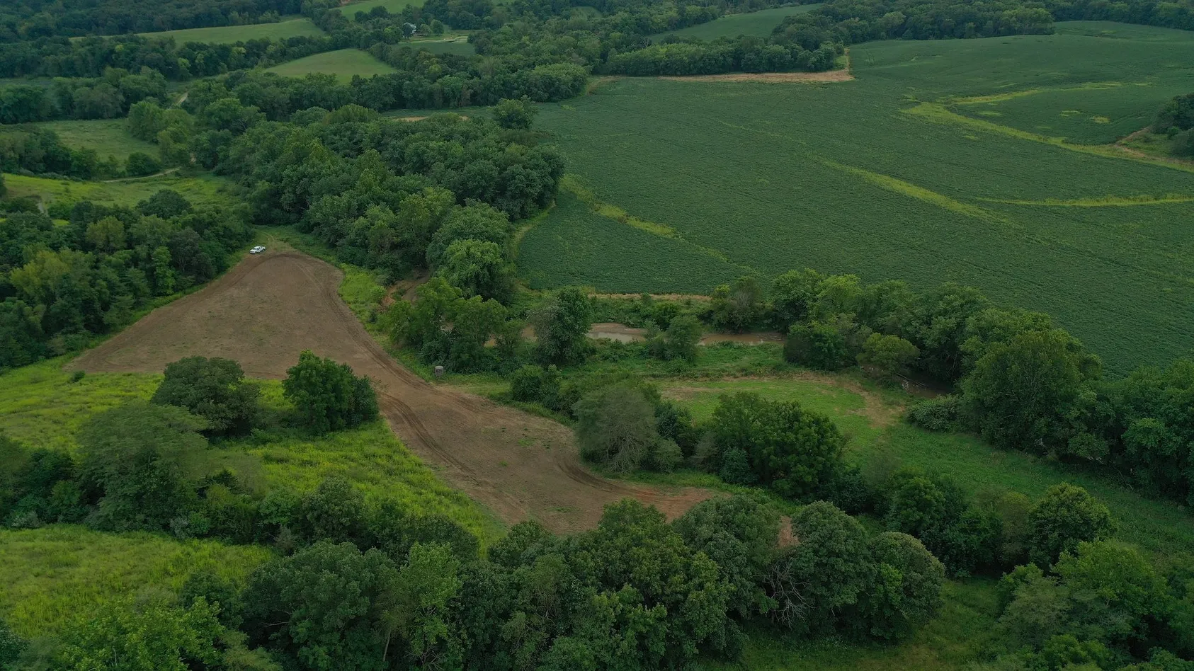 Aerial view of curved brown dirt trail through green agricultural land with scattered trees and woodland edges