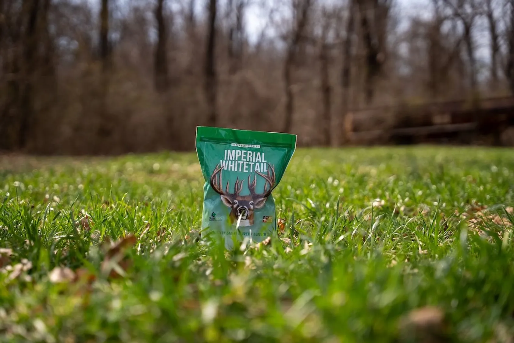 Imperial Whitetail seed bag with buck antler logo standing in green clover food plot with bare trees behind.