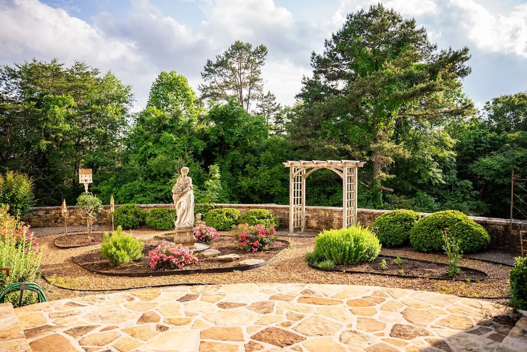 Formal garden with stone statue, pink flowers, wooden arbor, and stone wall bordered by evergreens.