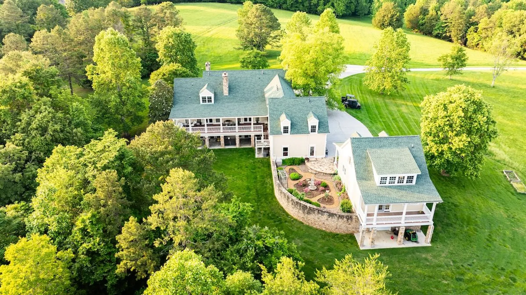 Aerial view of white two-story house with wraparound porch and detached garage surrounded by mature trees on lawn.