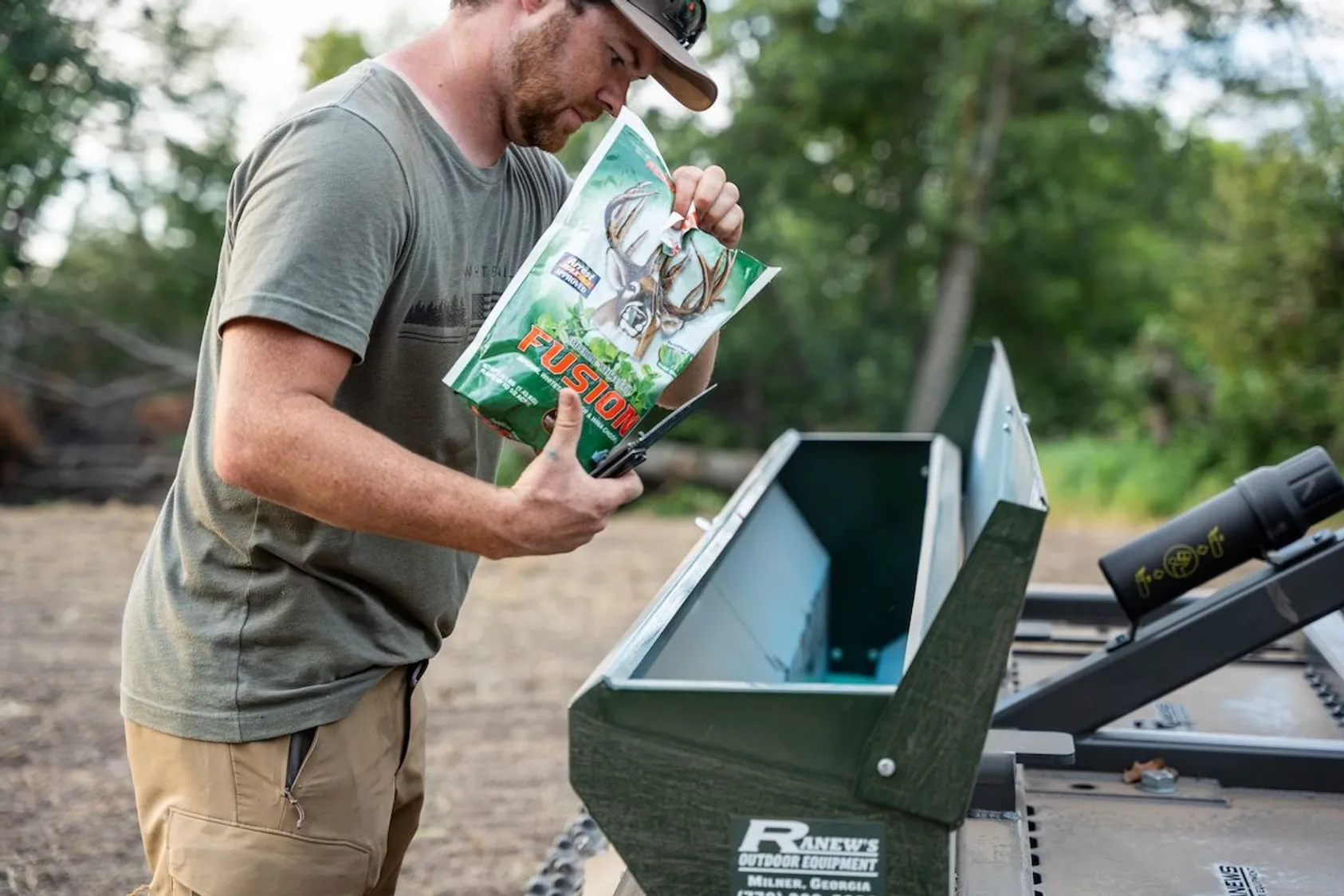 Man in cap and gray shirt pouring Fusion brand deer feed into metal feeder mounted on utility vehicle.