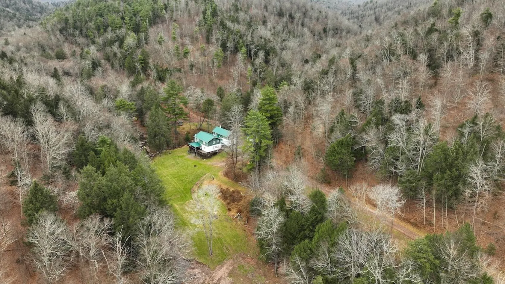 Aerial view of white house with teal roof in forest clearing surrounded by mixed bare and evergreen trees with dirt driveway.