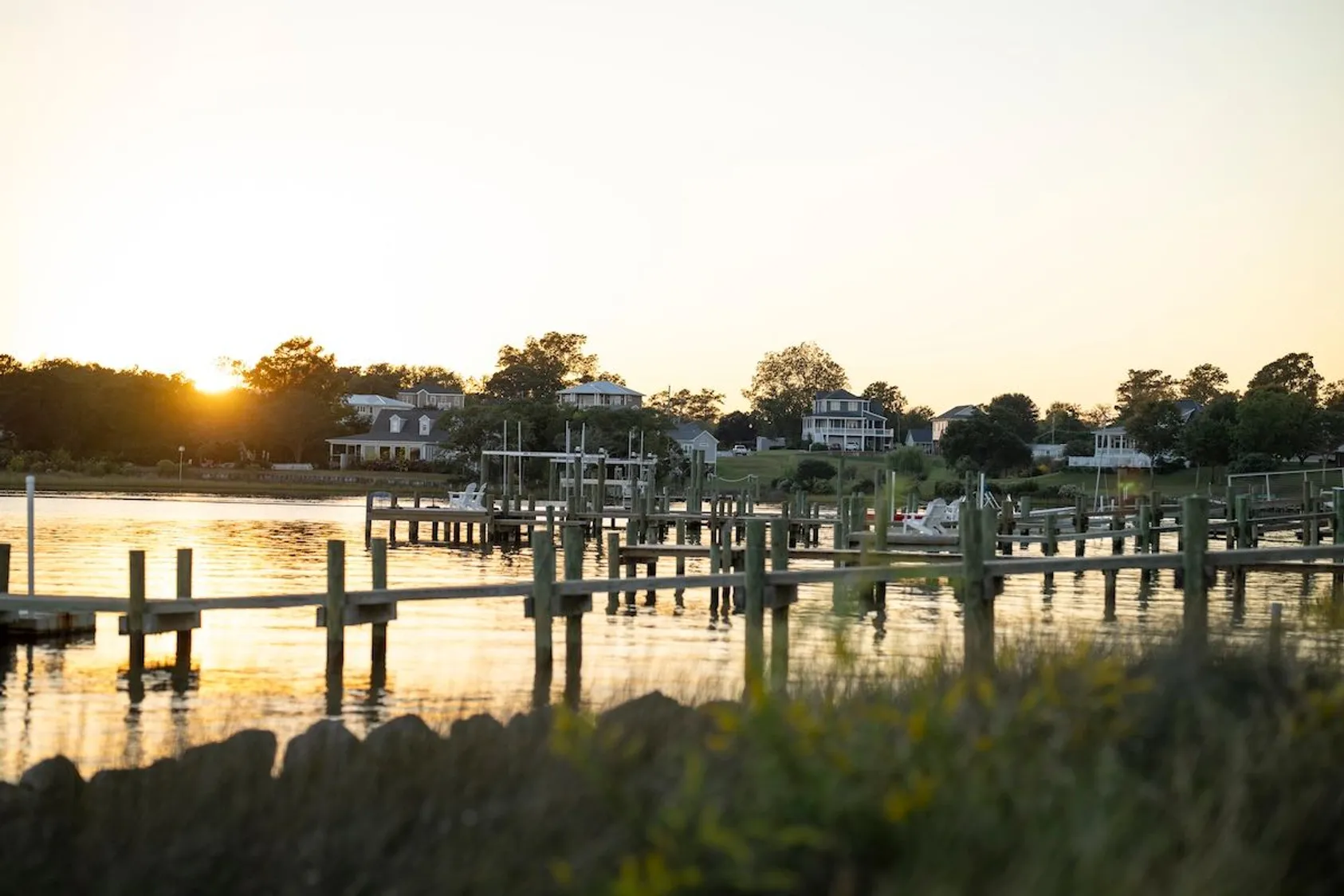 Marina at golden hour with multiple wooden boat docks and pilings reflected in calm water with waterfront homes and trees beyond.