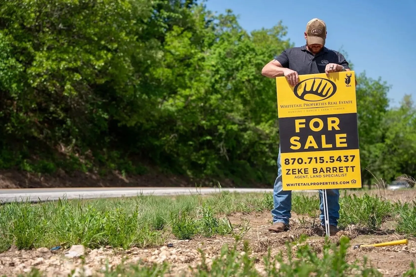 Man in cap and work clothes installing a yellow Whitetail Properties Real Estate For Sale sign along a roadside