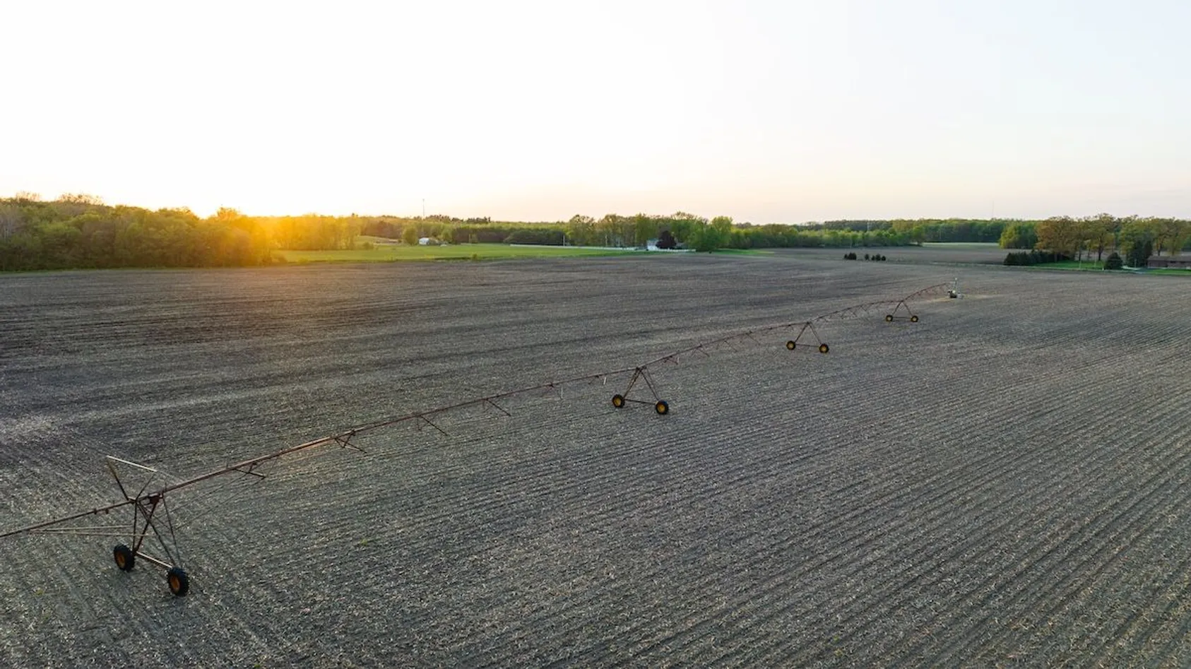 Pivot irrigation system stretching across tilled field at sunset with tree line in background.