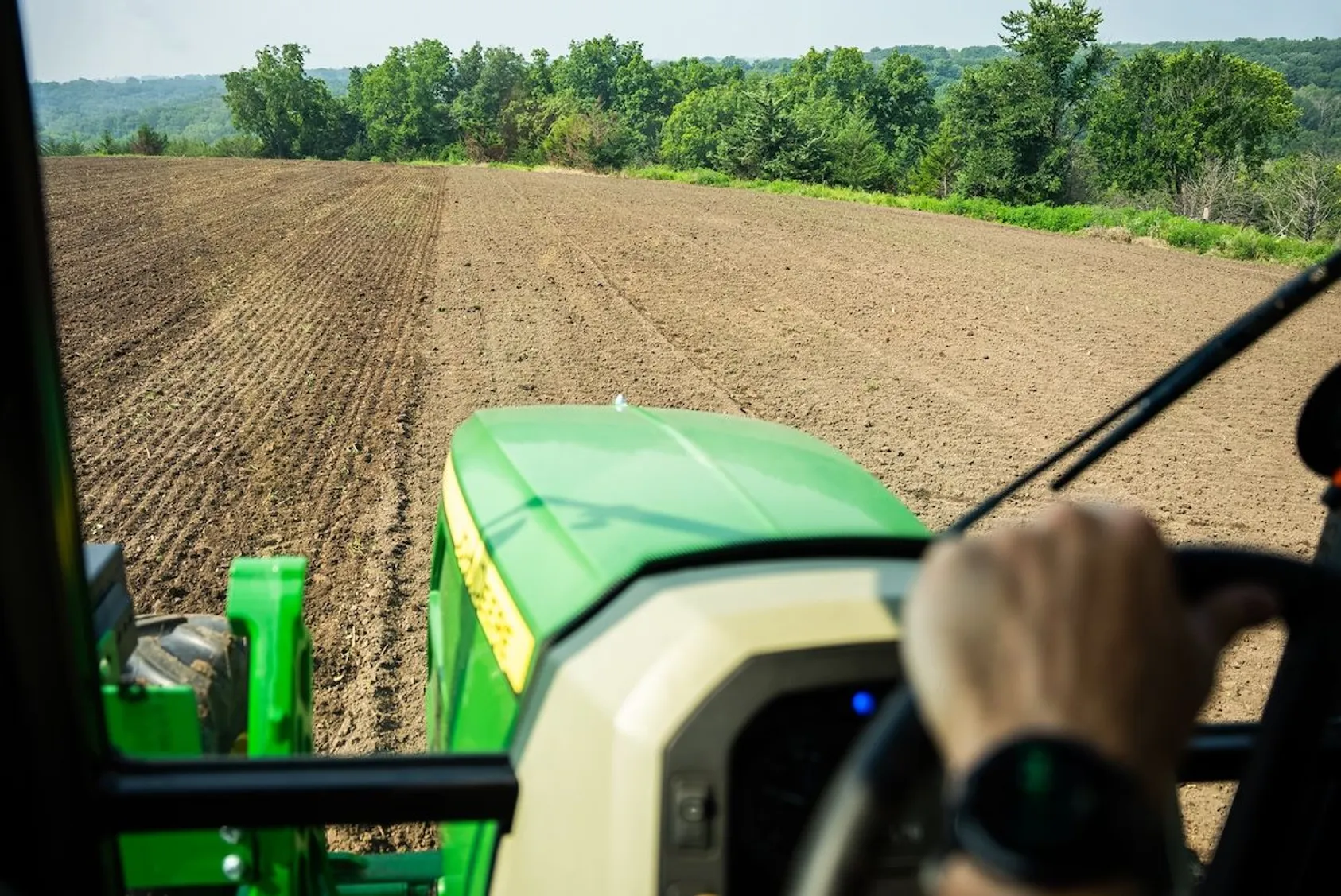 View from tractor cab showing freshly tilled brown field with tree line on horizon.