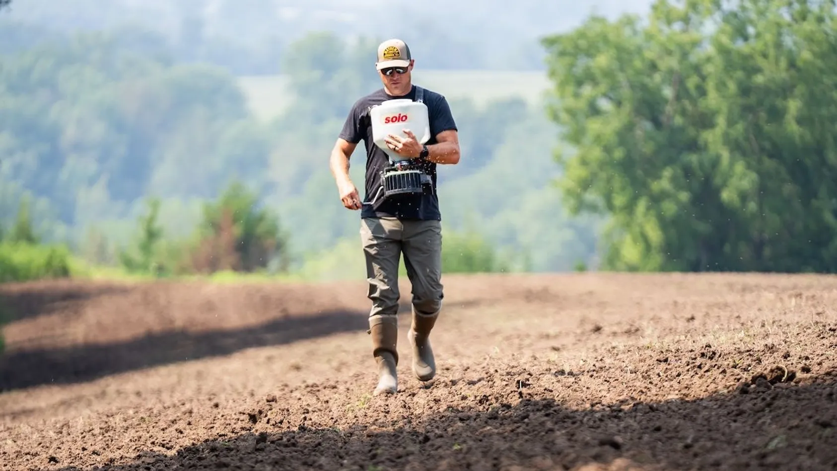 Man in cap and dark shirt using handheld broadcast spreader to seed tilled field with hills behind.