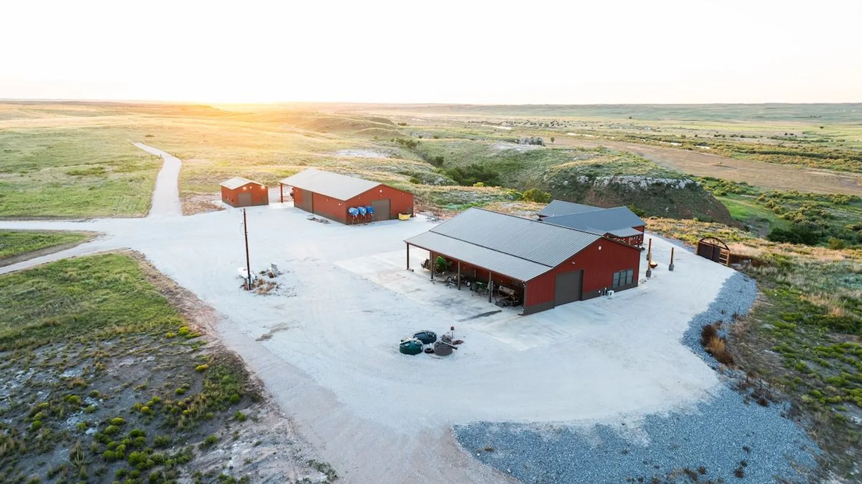 Aerial view of red barn buildings with metal roofs on concrete pad in rural prairie landscape at sunrise.