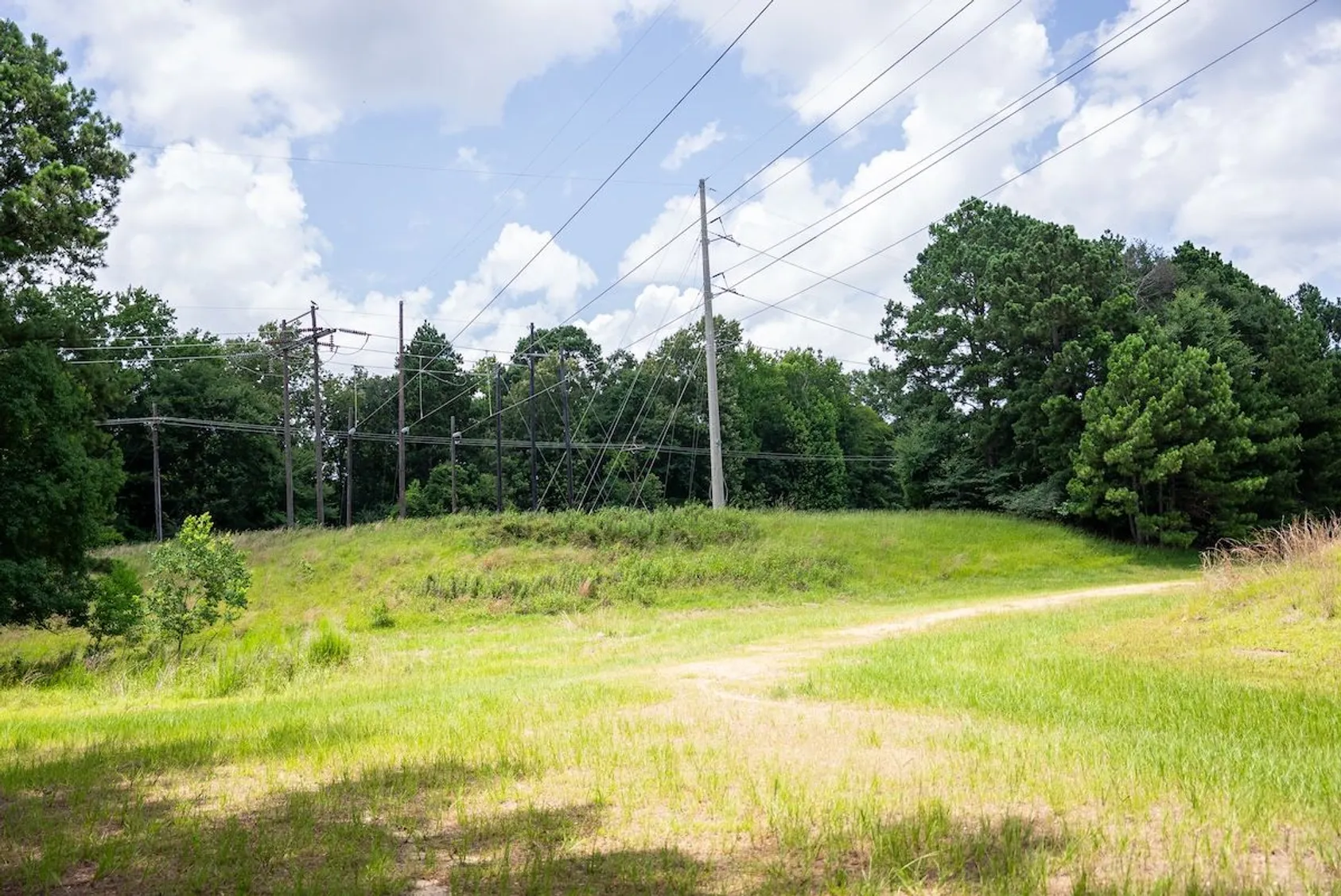 Power transmission lines and utility poles crossing grassy field with tree line under blue sky with clouds.