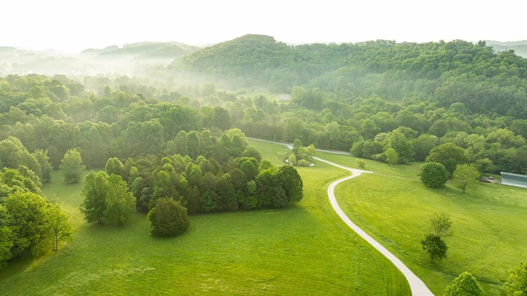 Misty morning aerial view of winding gravel road through green pasture surrounded by forested hills.