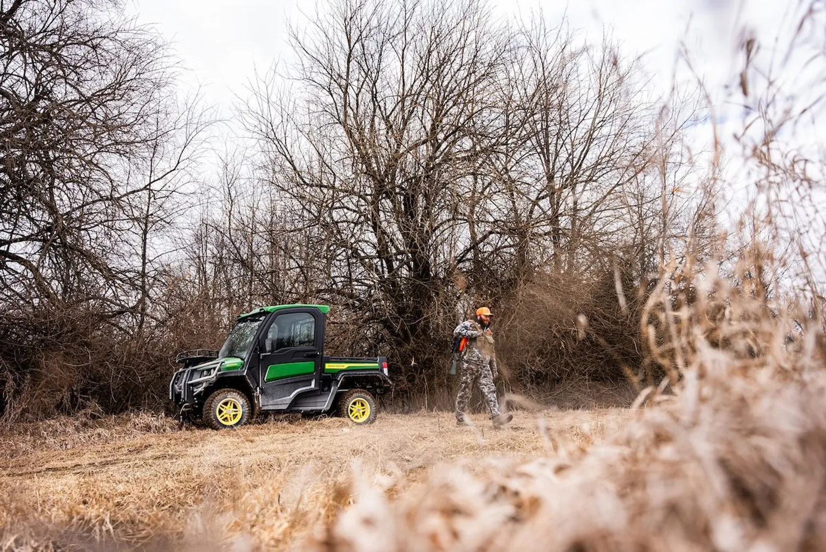 Hunter in orange cap and camouflage standing beside green John Deere utility vehicle in field near bare trees
