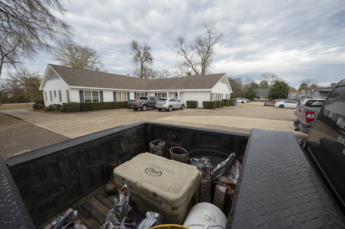 Pickup truck bed filled with tools and supplies in the foreground, with a white suburban house and parked cars under a cloudy sky in the background.