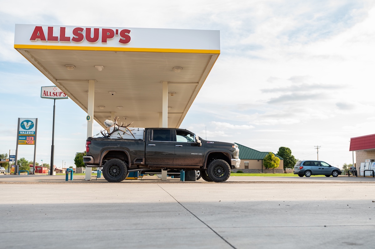 A black pickup truck with antlers in the back is parked under an Allsup's gas station canopy. The sky is bright with scattered clouds, and a blue car is in the background.