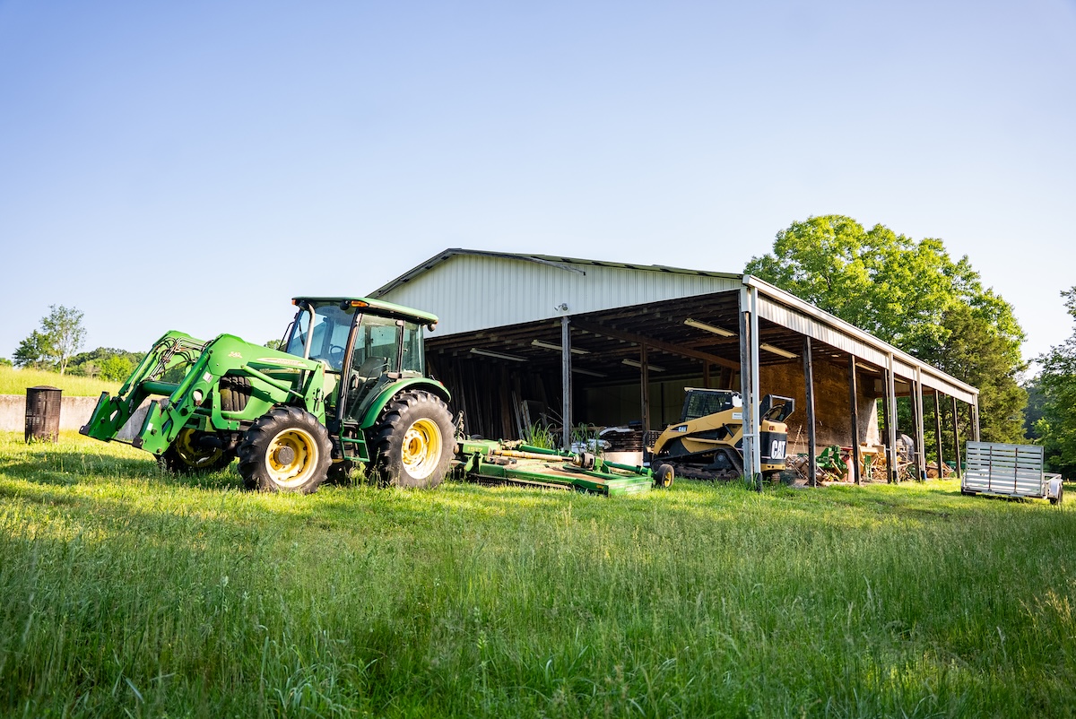 A green tractor is parked next to a large, open-sided barn filled with farming equipment. The scene is set in a lush, sunny field with trees in the background.