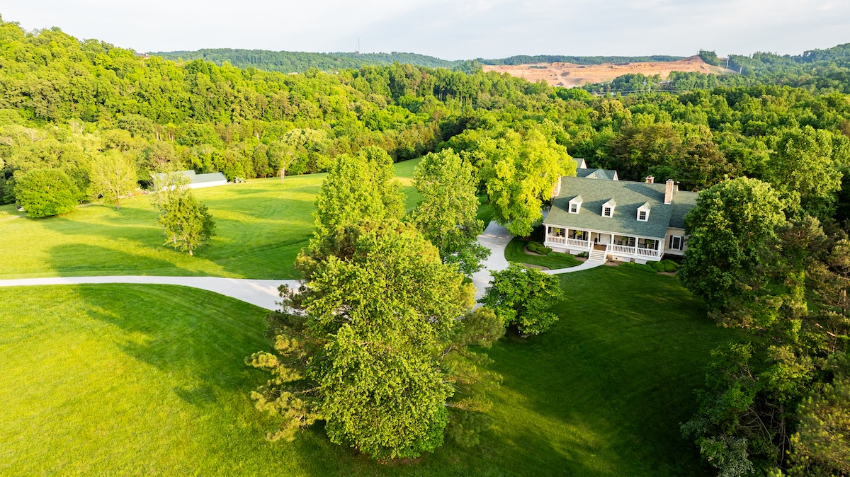 Aerial view of a large, elegant house with a green roof surrounded by lush trees and expansive lawns, set against a backdrop of hilly, wooded terrain.